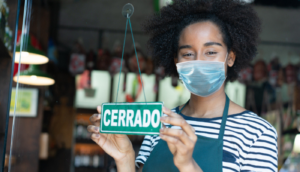 This stock image is of a Black female sales clerk at a delicatessen holding the closed sign wearing a protective face mask facing camera.