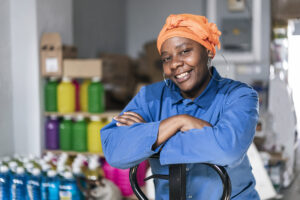 Mid adult Black woman with a uniform and headscarf smiling and sitting in a distribution warehouse.