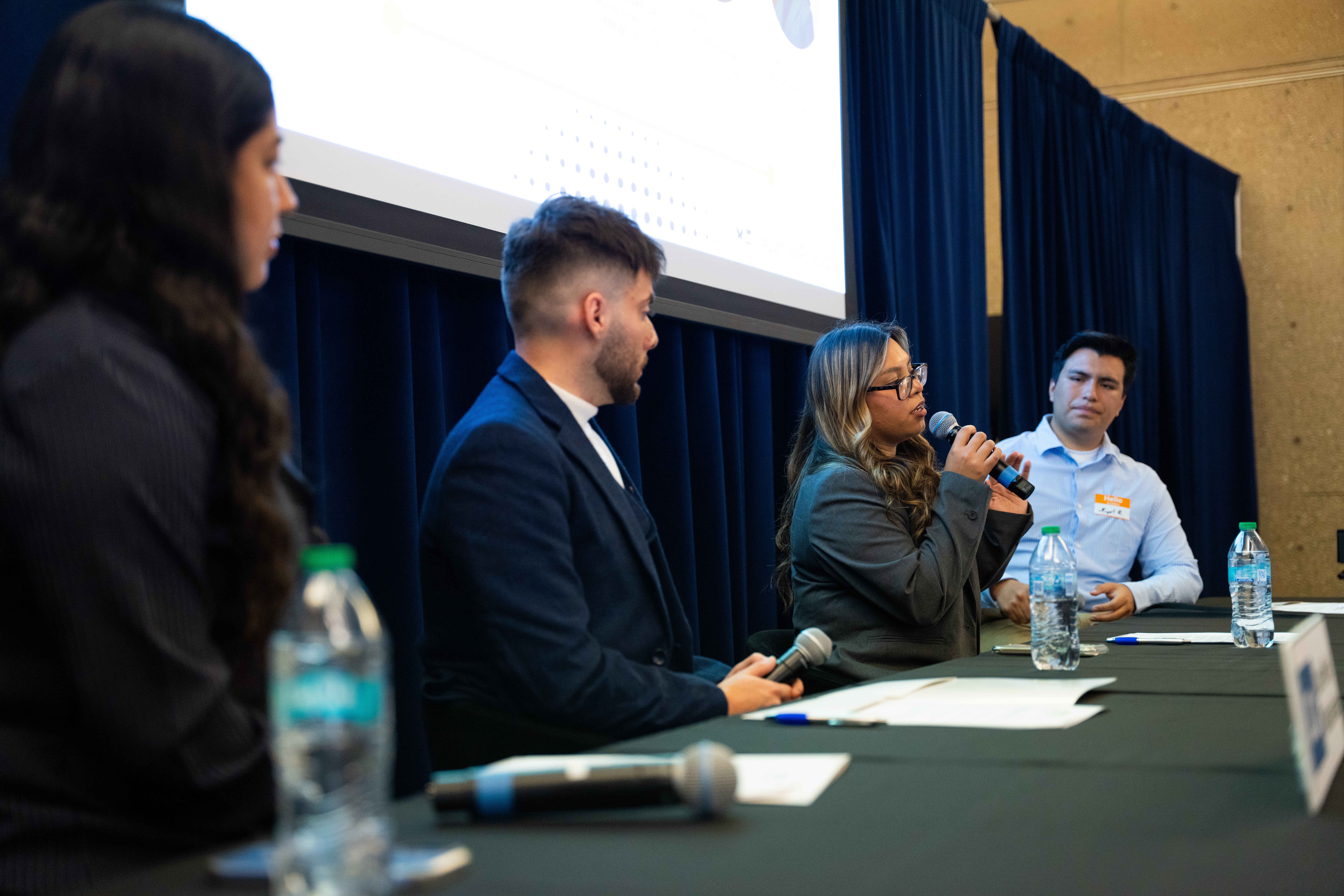 A young Latina woman speaks into a microphone, on a panel with two young Latino men and another young Latina.