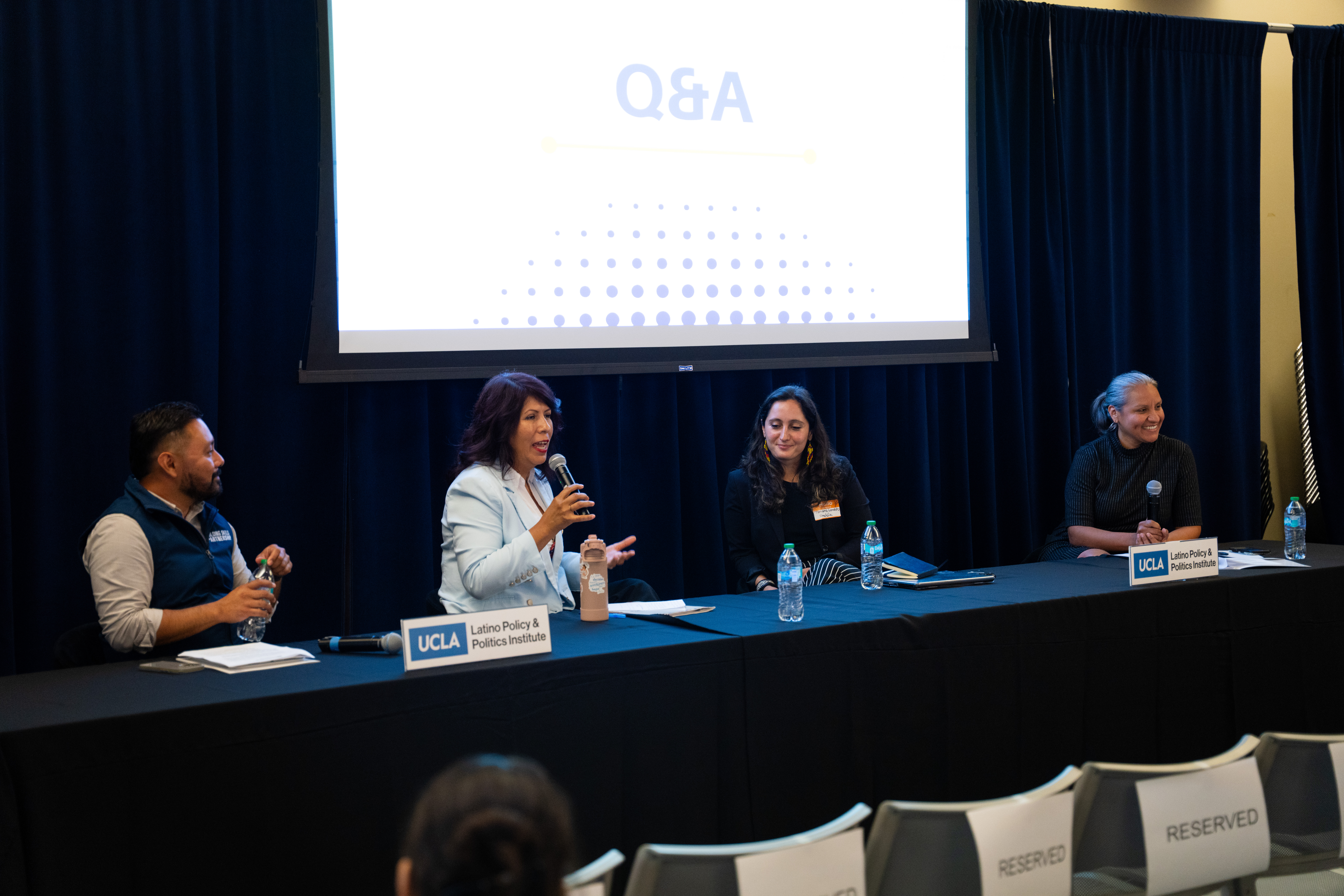 A middle-aged Latina woman speaks on a panel with other Latina women and one Latino man.