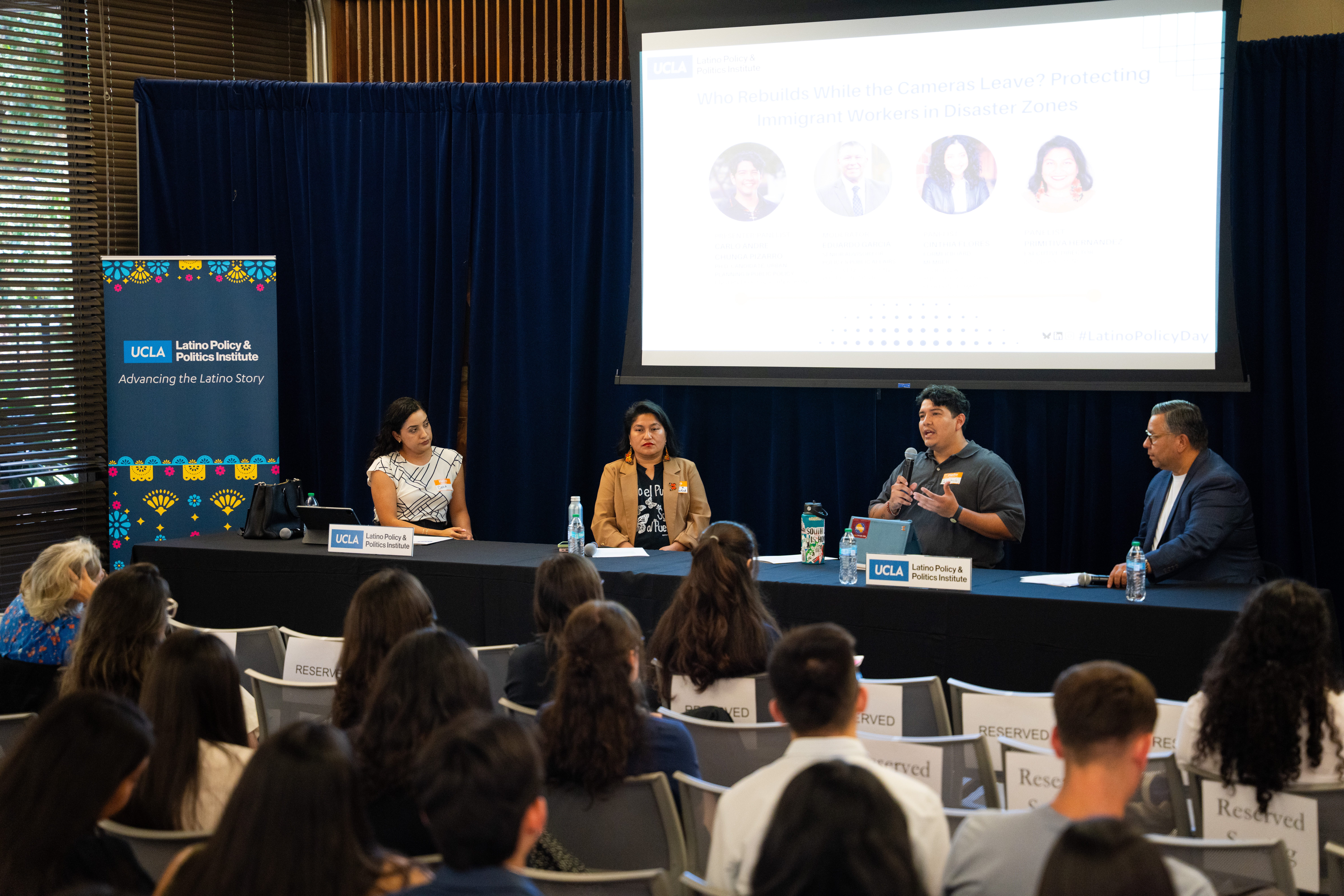 A young Latino man speaks into a microphone on a panel with two Latina women and a Latino man. An audience is seen in the foreground and a projector screen in the background.