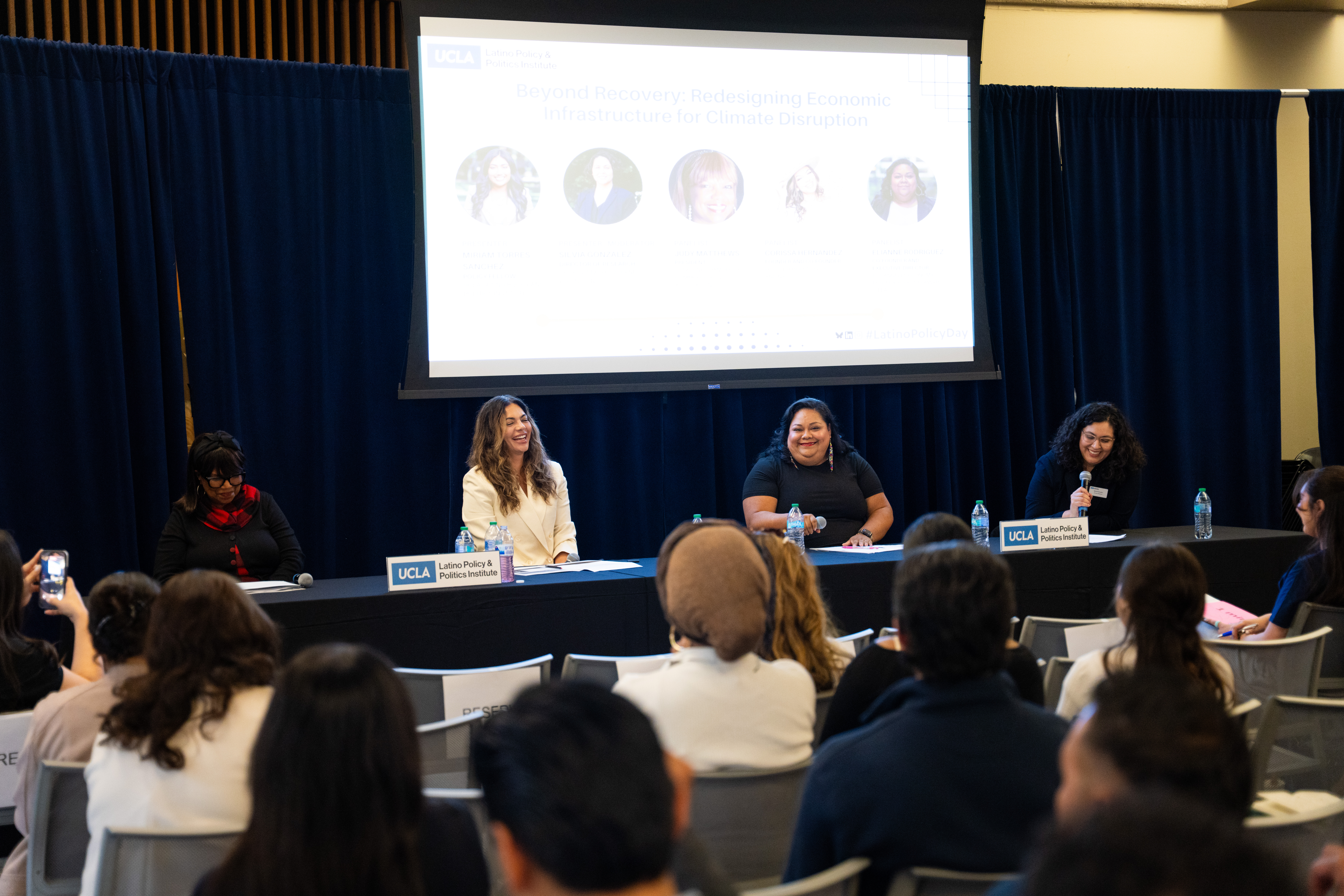 Three Latina women and one Black woman laugh on a panel. An audience is seen in the foreground and a projector screen in the background.